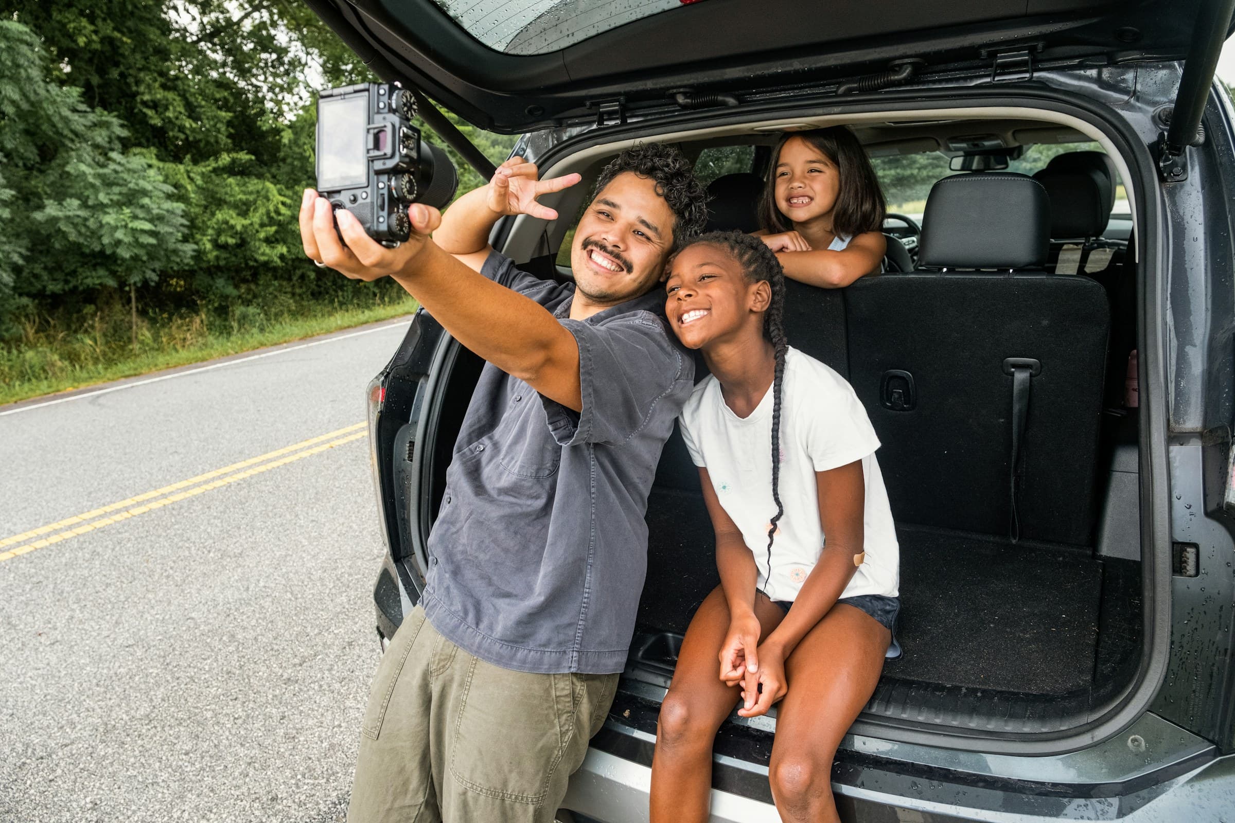 A father and his two daughters laughing together in the open boot of a people mover, parked at the roadside before setting off